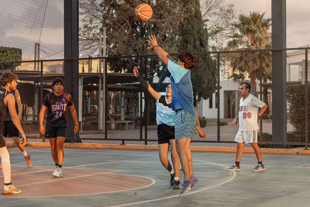Jogo de basquete ao ar livre com jogadores arremessando e defendendo em uma quadra de rua.
